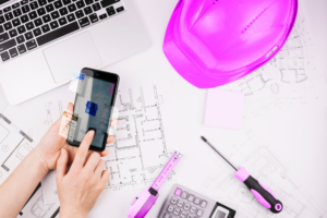 Construction contractor working at desk with a laptop, papers, helmet, tools, and calculator, using cell phone for Google Local Service Ads for Construction strategy.